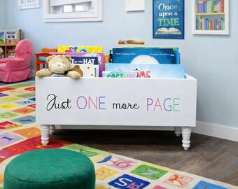 May include: White wooden bookshelf with the text "Just ONE more PAGE" in colorful letters. The bookshelf is filled with books and a stuffed teddy bear. The room has a colorful rug and a green ottoman.