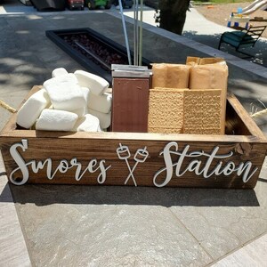 May include: A rectangular dark brown wooden crate with two jute rope handles, featuring the white 3D text "S'mores Station" with a marshmallow graphic. The crate is filled with white marshmallows, rectangular chocolate bars, and graham crackers.