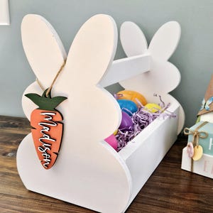 May include: White wooden bunny-shaped Easter basket with a handle. The basket is filled with colorful plastic eggs and purple shredded paper. A wooden carrot with the name "Madison" is hanging from the basket.