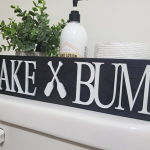 May include: A black wooden sign with white lettering that reads "LAKE X BUM" with crossed oars. The sign is placed on a white toilet. A bottle of hand lotion, a small plant, and a roll of toilet paper are on the shelf above the sign.