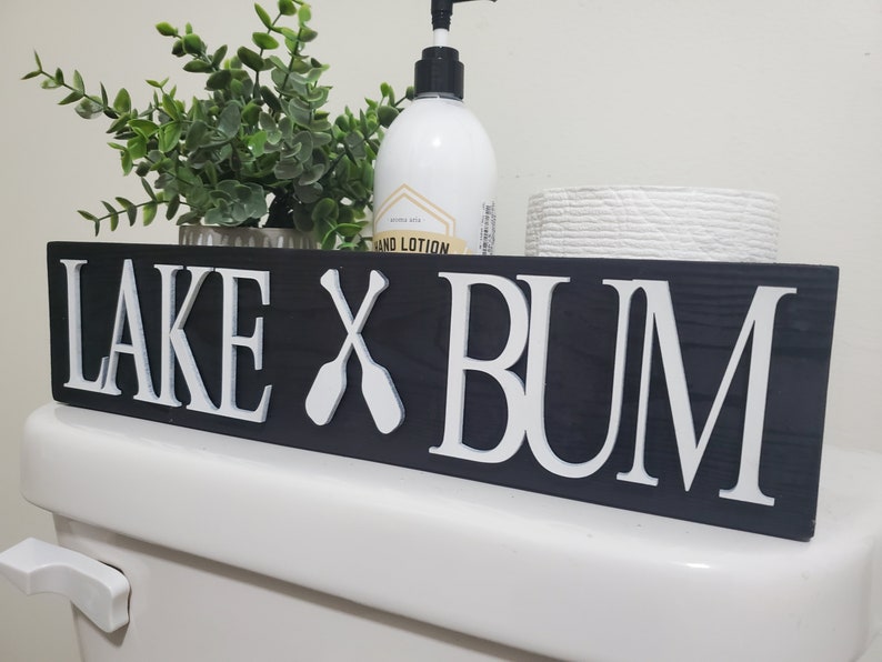 May include: A black wooden sign with white lettering that reads "LAKE X BUM" with crossed oars. The sign is placed on a white toilet. A bottle of hand lotion, a small plant, and a roll of toilet paper are on the shelf above the sign.
