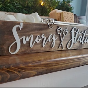 May include: A rectangular dark brown wooden box labeled S'mores Station in white 3D lettering with a graphic of two crossed marshmallows on sticks. The box contains white marshmallows and square graham crackers, with metal skewers resting inside.