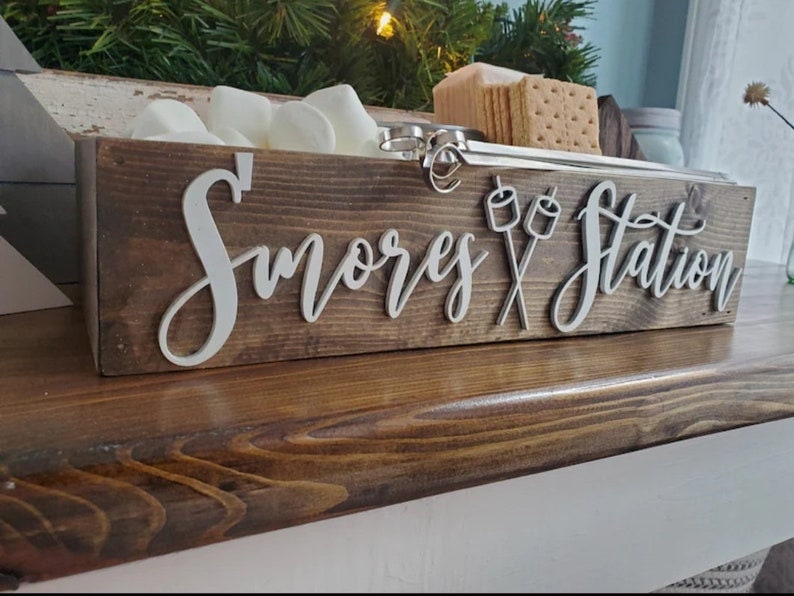 May include: A rectangular dark brown wooden box labeled S'mores Station in white 3D lettering with a graphic of two crossed marshmallows on sticks. The box contains white marshmallows and square graham crackers, with metal skewers resting inside.