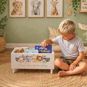 May include: A white wooden toy chest with a jungle animal design and the text "Just one more story." The chest is filled with books and toys. The child is reaching for a blue book. The chest is on a round jute rug.