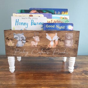 May include: Wooden book bin with white legs, holding children's books. The bin is stained brown and decorated with woodland animal illustrations. The text "Baby Colon's Books" is written on the front.