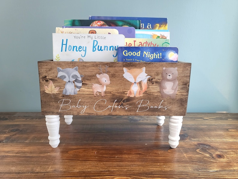 May include: Wooden book bin with white legs, holding children's books. The bin is stained brown and decorated with woodland animal illustrations. The text "Baby Colon's Books" is written on the front.