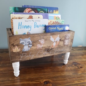 May include: Wooden bookshelf with white legs, holding children's books. The front of the shelf is decorated with woodland animal illustrations and the text "Baby [Name]'s Books". The books include titles like "You're My Little Honey Bunny" and "Good Night!"
