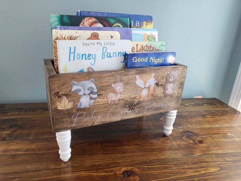 May include: Wooden bookshelf with white legs, holding children's books. The front of the shelf is decorated with woodland animal illustrations and the text "Baby [Name]'s Books". The books include titles like "You're My Little Honey Bunny" and "Good Night!"