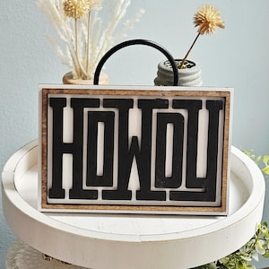 May include: A wooden sign with the word "HOWDY" in bold, black letters. The sign has a white background and a natural wood frame. A black handle arches over the top. The sign is on a white tray with decorative plants.