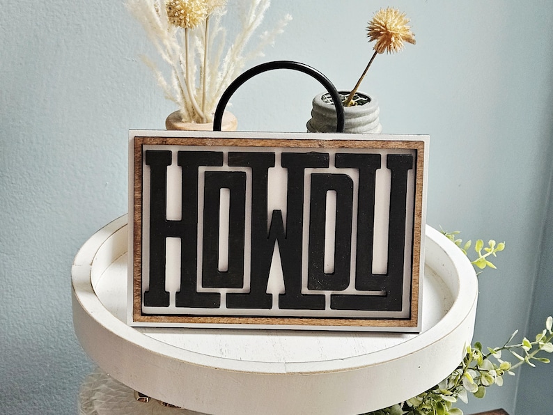 May include: A wooden sign with the word "HOWDY" in bold, black letters. The sign has a white background and a natural wood frame. A black handle arches over the top. The sign is on a white tray with decorative plants.