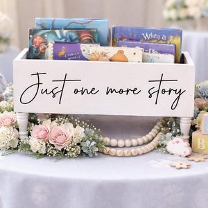 May include: A white wooden box with the text "Just one more story" holds children's books. The box is decorated with flowers, wooden beads, and blocks that spell "BABY". A gray stuffed elephant sits nearby.