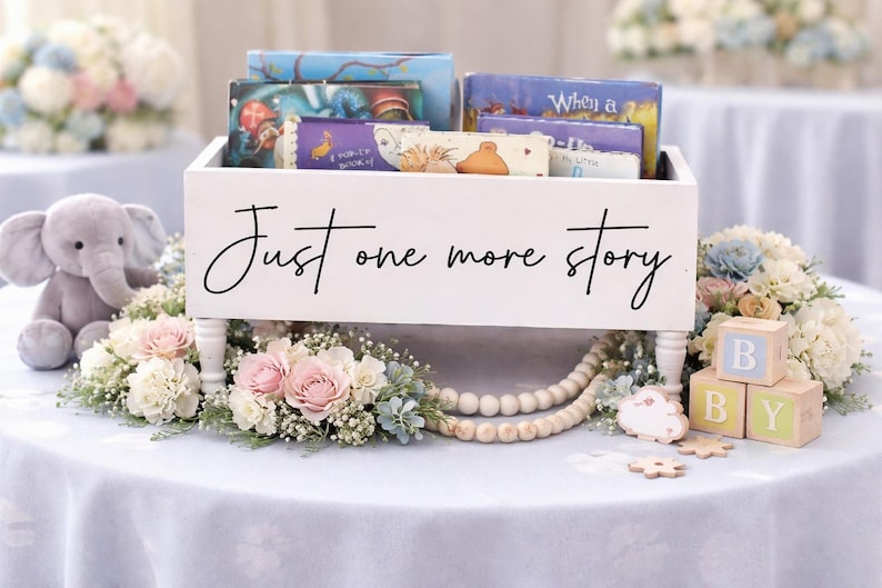 May include: A white wooden box with the text "Just one more story" holds children's books. The box is decorated with flowers, wooden beads, and blocks that spell "BABY". A gray stuffed elephant sits nearby.