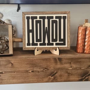 May include: A framed sign with the word "HOWDY" in bold, black letters. The sign is displayed on a wooden shelf, alongside a decorative pumpkin and a clock. The sign is supported by a wooden stand.