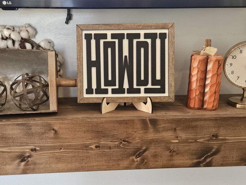 May include: A framed sign with the word "HOWDY" in bold, black letters. The sign is displayed on a wooden shelf, alongside a decorative pumpkin and a clock. The sign is supported by a wooden stand.