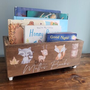 May include: A wooden book bin with a rustic finish, filled with children's books. The bin is decorated with woodland animal illustrations and the name "Mattis Books". It sits on small white wheels.