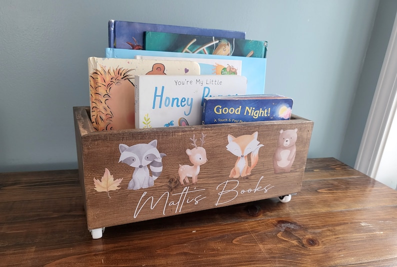 May include: A wooden book bin with a rustic finish, filled with children's books. The bin is decorated with woodland animal illustrations and the name "Mattis Books". It sits on small white wheels.
