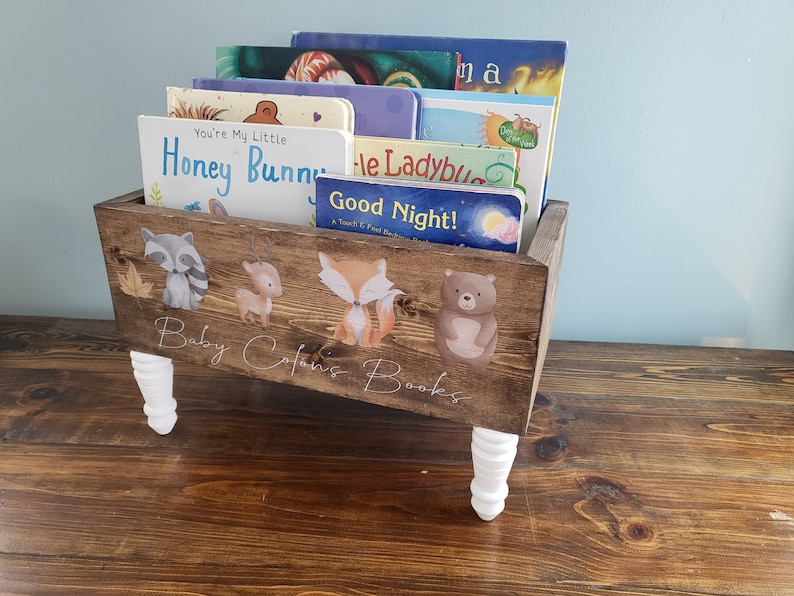 May include: A wooden bookshelf with a dark brown finish, filled with children's books. The front of the shelf is decorated with woodland animal illustrations and the text "Baby Colors Books". The shelf has white, decorative legs.