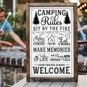 May include: A black and white sign with the text "Camping Rules" and a list of rules for camping, including "Sit by the fire", "Relax and unwind", "Eat s'mores", "Make memories", "Wake up late", "Keep the drinks cold", and "Good friends always". The sign is framed in wood.