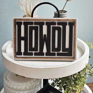 May include: A rectangular wooden sign with the word "HOWDY" in bold, black letters. The sign has a white background and a dark brown wooden frame. A black handle arches over the top. Decorative plants and a small house are in the background.