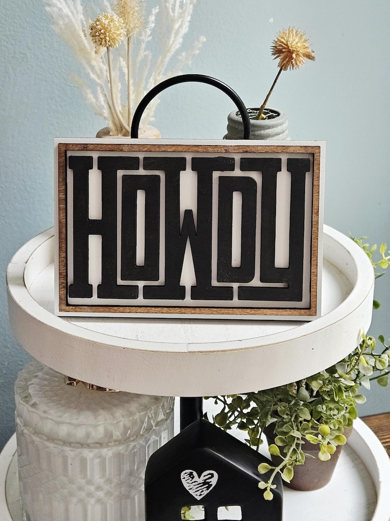 May include: A rectangular wooden sign with the word "HOWDY" in bold, black letters. The sign has a white background and a dark brown wooden frame. A black handle arches over the top. Decorative plants and a small house are in the background.