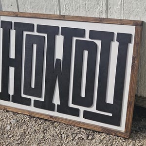 May include: A rectangular wooden sign with the word "HOWDY" in large, black, block letters. The letters are raised against a white background, framed by a dark brown wooden border. The sign is leaning against a white wall.