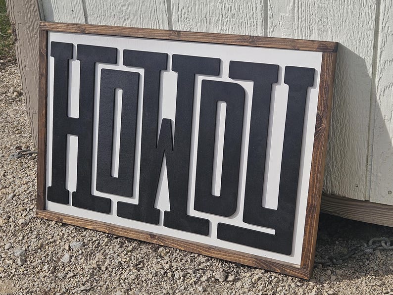 May include: A rectangular wooden sign with the word "HOWDY" in large, black, block letters. The letters are raised against a white background, framed by a dark brown wooden border. The sign is leaning against a white wall.
