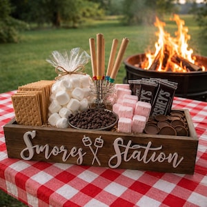 Könnte beinhalten: Ein rechteckiges Holztablett mit der Aufschrift S'mores Station und einer Marshmallow-Grafik auf der Vorderseite. Es enthält Graham-Cracker, eine Tüte weiße Marshmallows, eine Schüssel mit Schokoladenstückchen, rosa Marshmallows und Röstspieße.