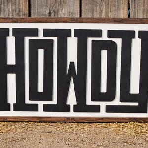 May include: A rectangular wooden sign with the word "HOWDY" in bold, black letters. The letters are set against a white background and framed by a dark brown wooden border. The sign is displayed outdoors.