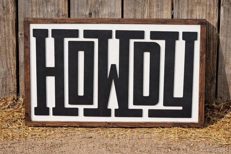 May include: A rectangular wooden sign with the word "HOWDY" in bold, black letters. The letters are set against a white background and framed by a dark brown wooden border. The sign is displayed outdoors.