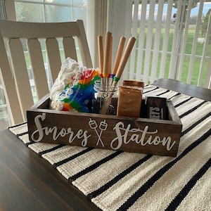 May include: A rectangular dark brown wooden box with the white text S'mores Station and a graphic of two marshmallows on sticks. Inside are a clear glass jar with four metal roasting skewers, a bag of marshmallows, and chocolate bars.