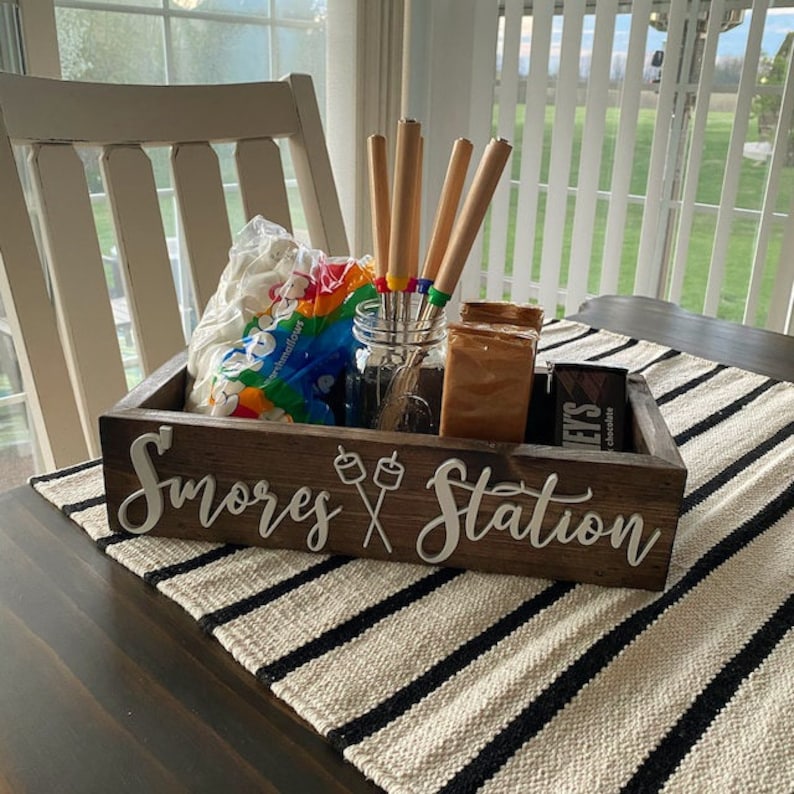 May include: A rectangular dark brown wooden box with the white text S'mores Station and a graphic of two marshmallows on sticks. Inside are a clear glass jar with four metal roasting skewers, a bag of marshmallows, and chocolate bars.