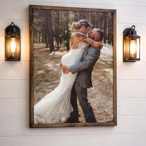 May include: A framed wedding photo shows a couple embracing in a forest setting. The bride wears a white lace gown, and the groom is in a gray suit. Two black sconces with Edison bulbs flank the framed photo on a white shiplap wall.