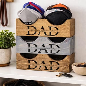 May include: Wooden hat organizer with three compartments, each labeled "DAD" in black script. The top compartment holds baseball caps in various colors. A small plant and other accessories are visible.