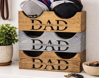May include: Wooden hat organizer with three compartments, each labeled "DAD" in black script. The top compartment holds baseball caps in various colors. The organizer is on a white shelf with a woven basket and shoes.