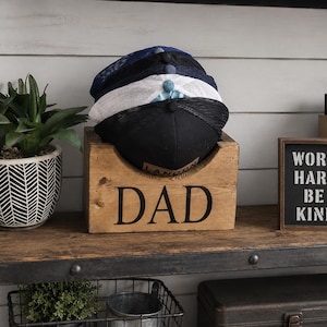 May include: A wooden box with the word "DAD" in large black letters holds several baseball caps. The caps are black, white, and navy blue. A small framed sign reads "WORK HARD BE KIND". A potted plant sits to the left.