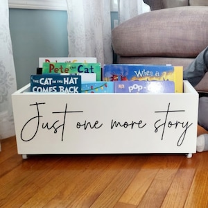 May include: A white wooden book bin on wheels, filled with children's books. The bin has the phrase "Just one more story" in black script. The books include "Pete the Cat" and "The Cat in the Hat Comes Back".