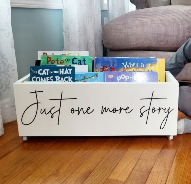 May include: A white wooden book bin on wheels, filled with children's books. The bin has the phrase "Just one more story" in black script. The books include "Pete the Cat" and "The Cat in the Hat Comes Back".