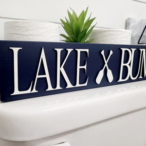 May include: A navy blue wooden sign with the words "LAKE X BUM" in white, with two white paddles replacing the "X". The sign is placed on a white toilet. A small green plant is visible.
