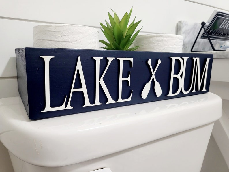 May include: A navy blue wooden sign with the words "LAKE X BUM" in white, with two white paddles replacing the "X". The sign is placed on a white toilet. A small green plant is visible.