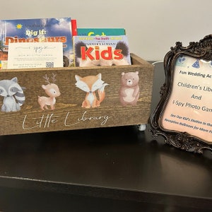May include: A wooden "Little Library" box with animal decals, holding children's books. The box features a raccoon, deer, fox, and bear. A sign in a decorative frame reads "Fun Wedding Activities" and "I-Spy Photo Game."