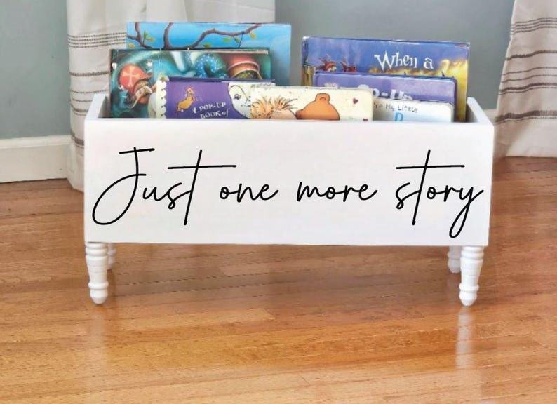 May include: A white wooden book bin with four short, turned legs. The bin is filled with colorful children's books. The words "Just one more story" are written in black script across the front of the bin.