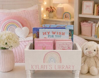 May include: A white wooden book bin with four short legs, featuring a pastel rainbow graphic and the text KYLAN'S LIBRARY printed on the front. The bin is filled with several children's books, including titles like MY WISH FOR YOU.
