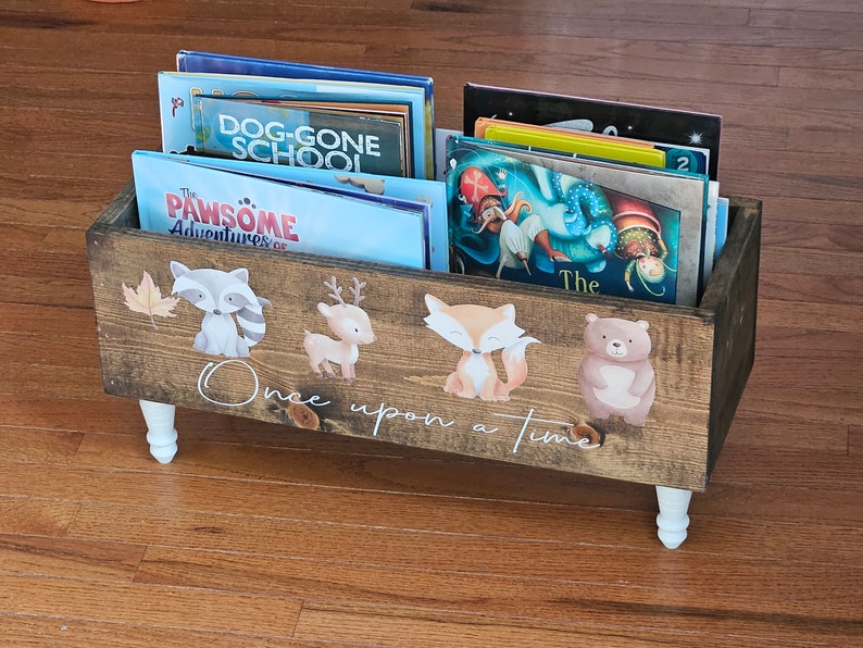 May include: Wooden book bin with white legs, filled with children's books. The front of the bin is decorated with woodland animal decals and the words "Once upon a time". The bin is made of stained wood.