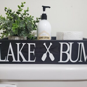 May include: A black wooden toilet tray with white lettering that reads "LAKE X BUM" and two crossed oars. The tray holds a small green plant, a hand lotion bottle, and a roll of toilet paper.
