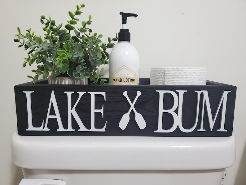 May include: A black wooden toilet tray with white lettering that reads "LAKE X BUM" and two crossed oars. The tray holds a small green plant, a hand lotion bottle, and a roll of toilet paper.