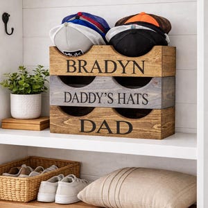 May include: A wooden hat organizer with three sections, each labeled with text. The top section says "BRADYN", the middle says "DADDY'S HATS", and the bottom says "DAD". Several baseball caps are visible in the top section. The organizer is on a shelf.