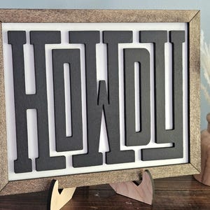 May include: Wooden framed sign with the word "HOWDY" in black, block letters. The sign is displayed on a wooden easel. A vase with dried flowers is to the right of the sign.
