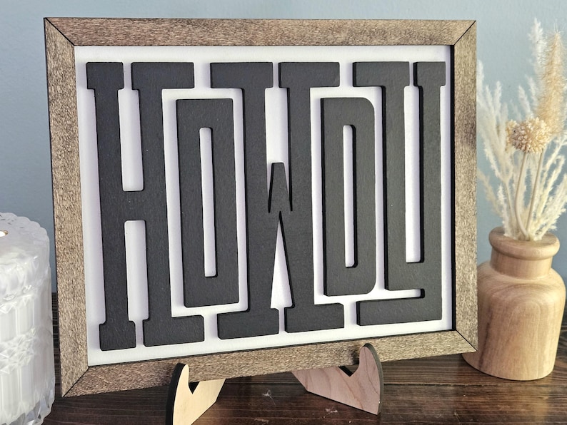 May include: Wooden framed sign with the word "HOWDY" in black, block letters. The sign is displayed on a wooden easel. A vase with dried flowers is to the right of the sign.