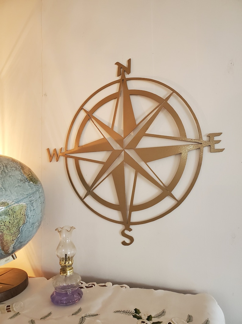 May include: A gold-colored metal compass rose wall decor. The compass rose features directional indicators: North, South, East, and West. The wall decor is mounted on a white wall. A globe and a small oil lamp are in the foreground.