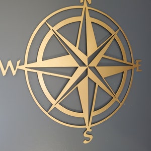 May include: A gold-colored compass rose wall decor. The compass features the cardinal directions: North, South, East, and West, with a star-shaped design in the center. The compass is mounted on a gray wall.